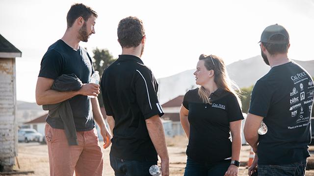 Four people stand outside in casual clothes, two holding water bottles, with buildings and hills in the background.