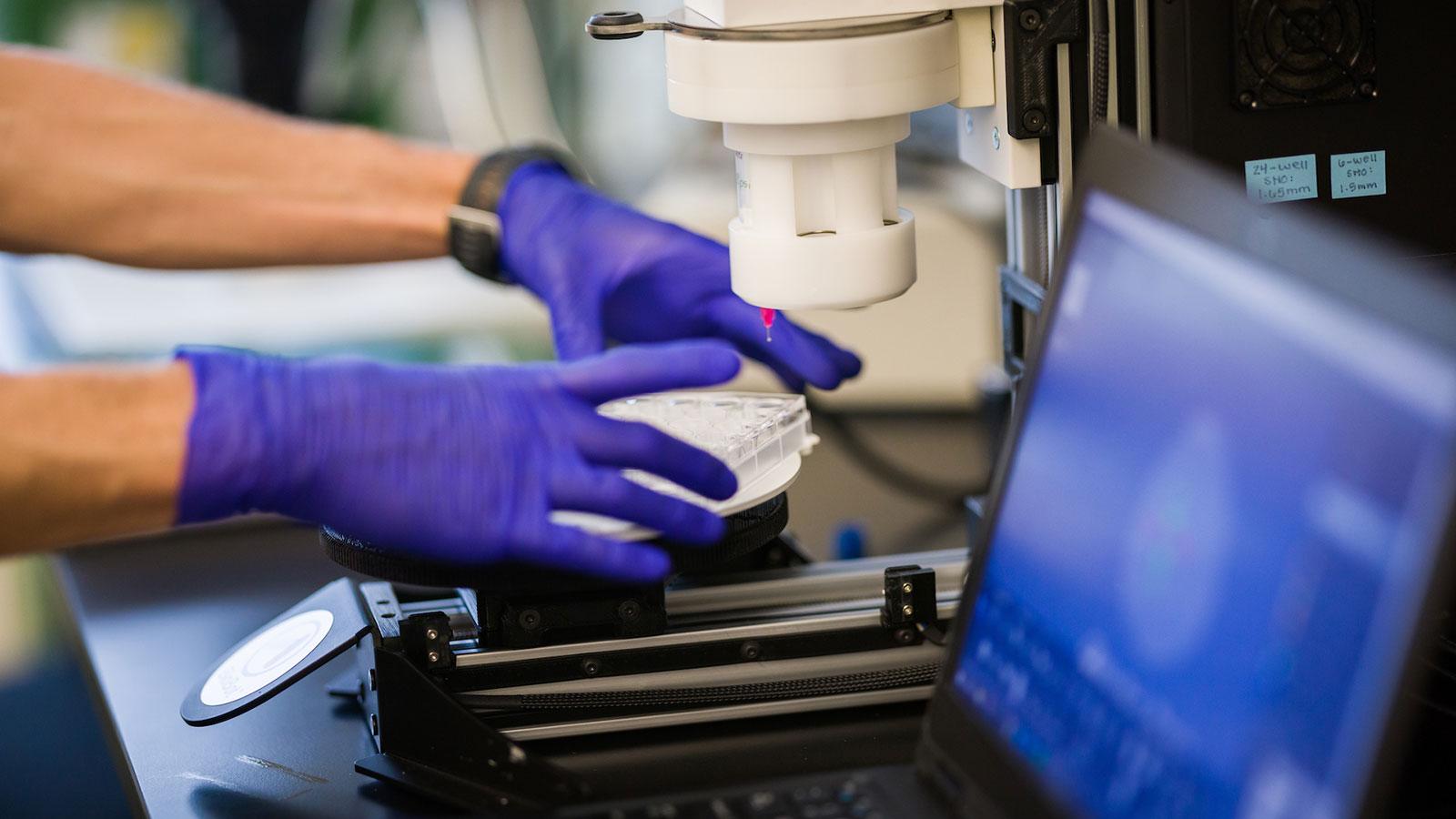 Gloved hands operate a laboratory instrument dispensing liquid into a multi-well plate during a biomedical engineering experiment.