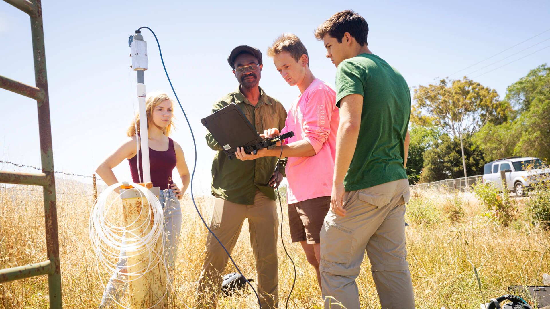 Students and instructor conducting outdoor field research using monitoring equipment in a grassy landscape.