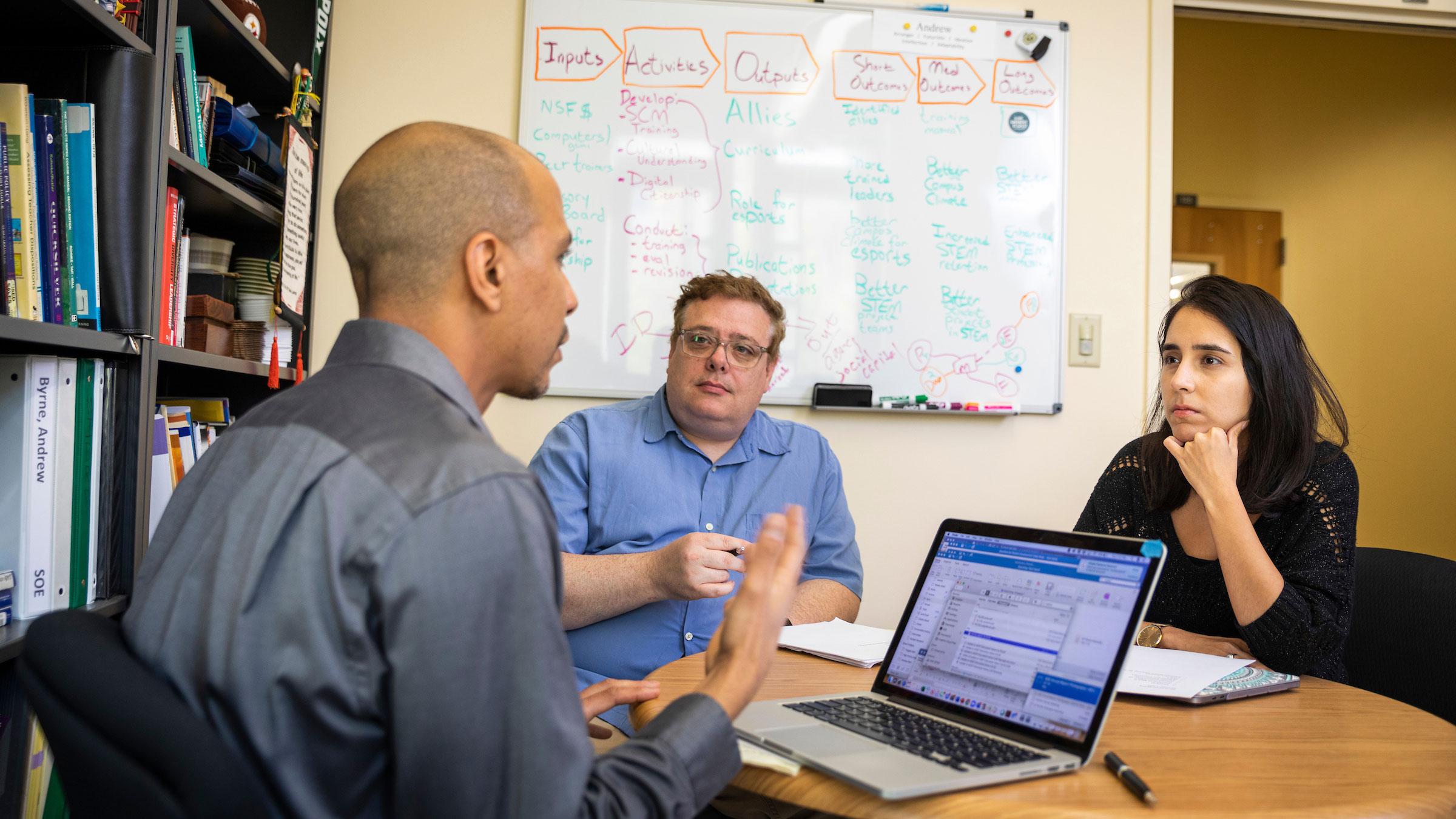 Three professionals seated at a table in discussion with a laptop and notes in an office setting.