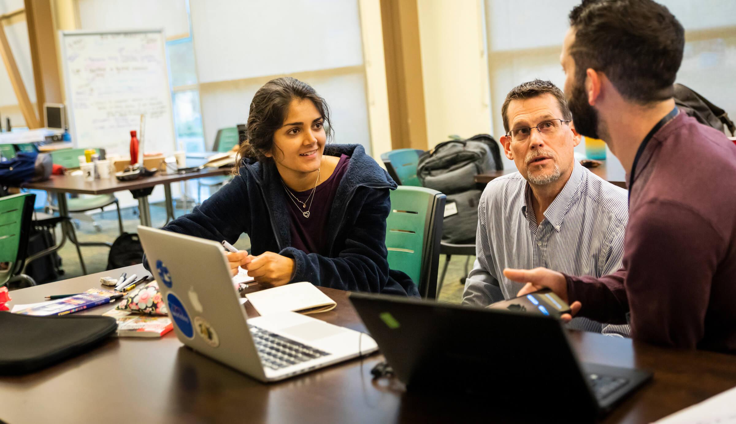 Three people engaged in a discussion around a table with laptops and notebooks in a casual workspace.