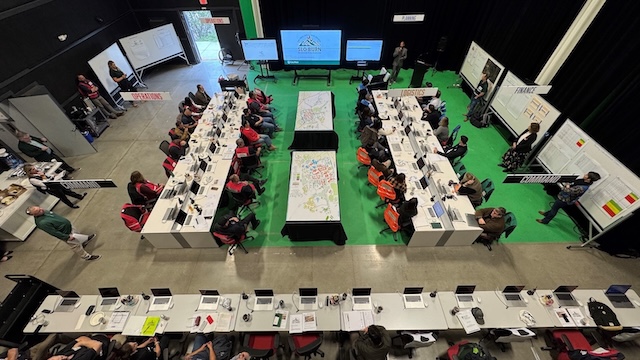 An overhead view of Cal Poly’s SLO BURN emergency simulation exercise, showing teams seated at long tables with laptops and maps in an operations room setup labeled with sections such as Command, Operations, Logistics, Planning, and Finance.